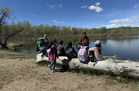 School children sitting on a log in front of a beautiful pond, listening intently to a Natural Areas staff member teaching them.